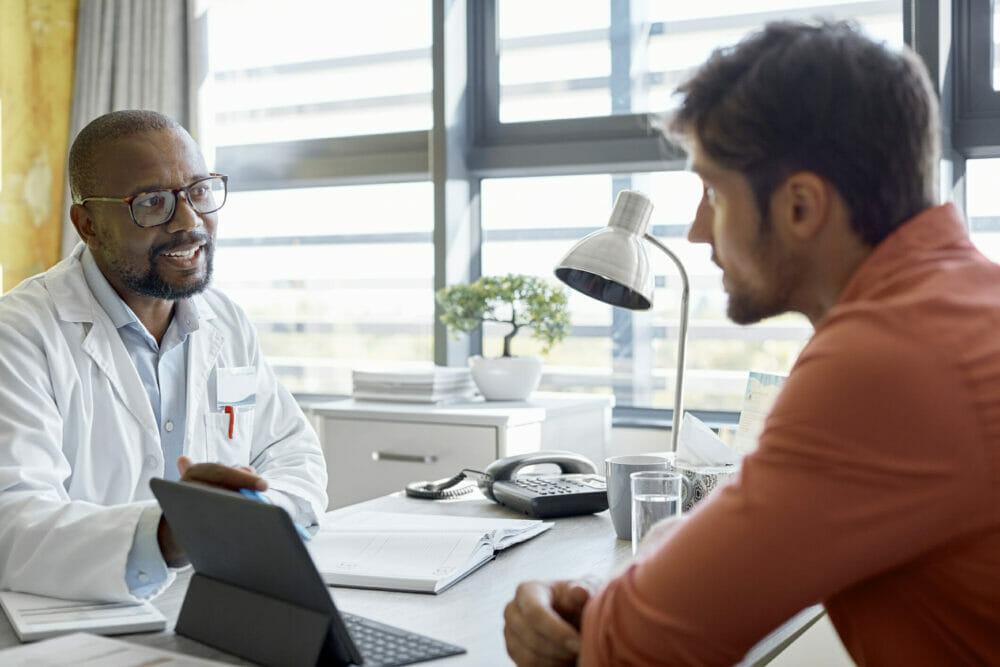 Male-doctor-and-young-male-patient-sitting-at-desk-in-a-clinic-having-a-discussion-while-using-a-digital-tablet.