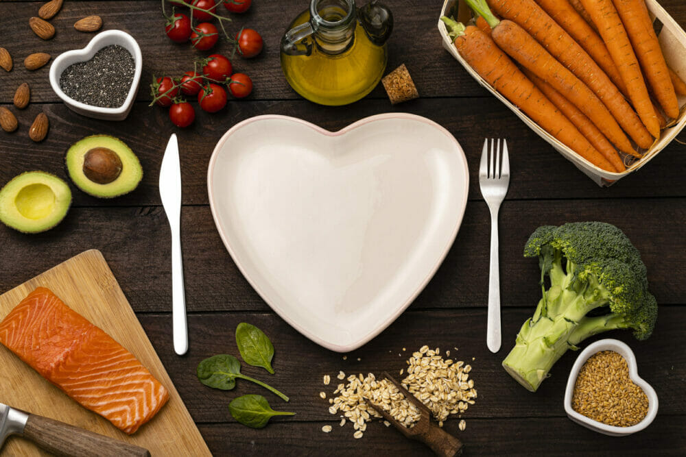 Top-view-of-a-heart shaped-plate-surrounded-by-a-knife-and-a-fork-and-some-healthy-food-such-as-a- salmon-fillet,-broccoli,-oat-flakes,-chia-and-flax-seeds,-cherry-tomatoes,-carrots,-almond,-avocado,-spinach-and-olive-oil.